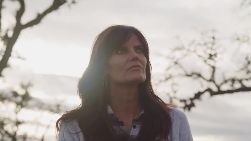 Middle aged woman with hoop earrings and wavy hair faces the camera without eye contact on a cloudy day