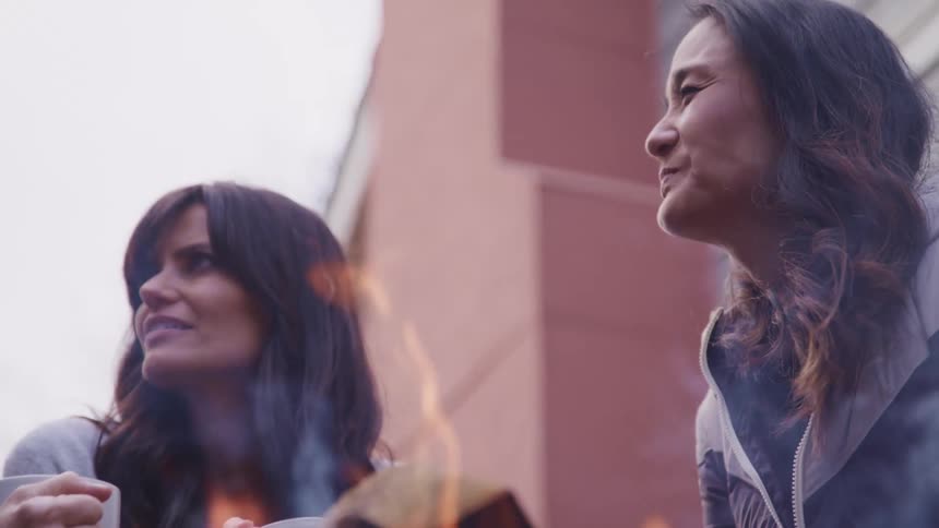Middle aged women chat with one another while sitting around a bonfire and holding coffee mugs