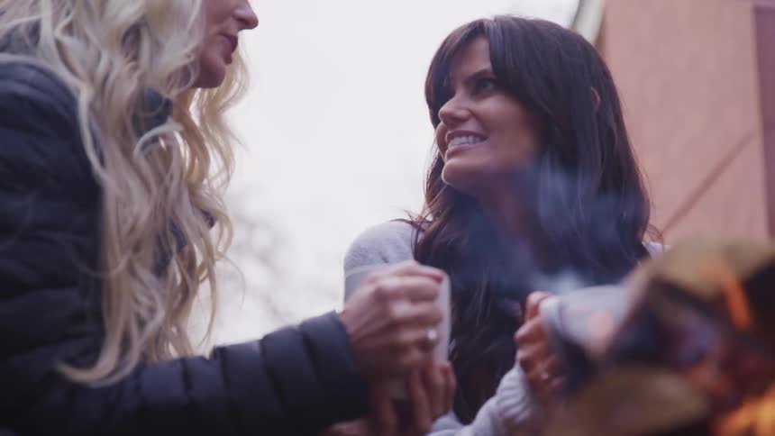 Two women sitting next to a fire outside a home talk while holding coffee mugs
