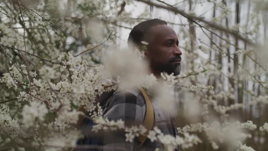 Side view of a black man with a beard a checkered shirt and a backpack standing under flowering trees