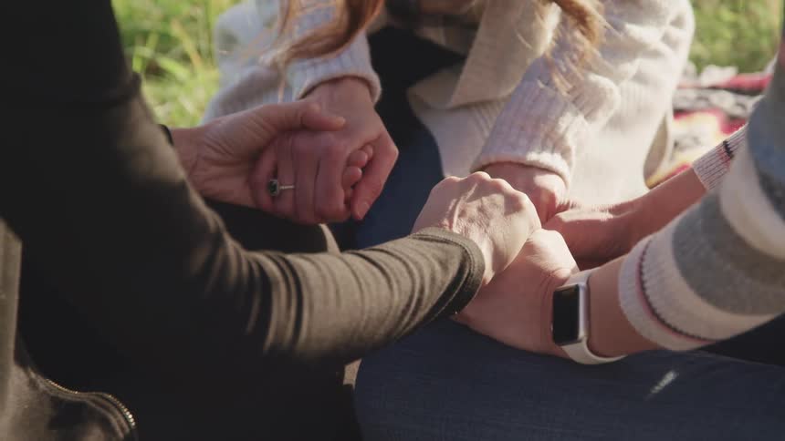 Three people hold hands while sitting in a field facing one another