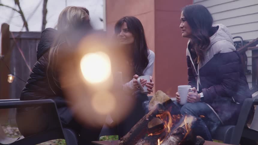 Three middle aged woman sit around a campfire in the backyard of a house