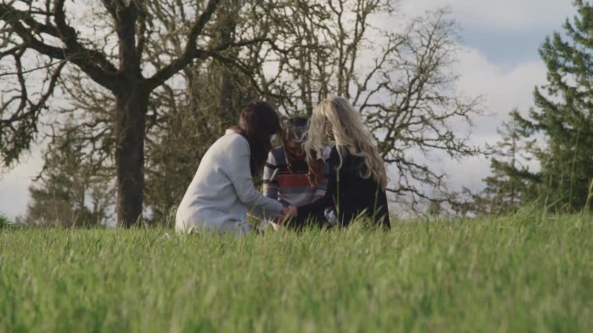 A trio of women sit in a circle holding hands with their heads bowed in a field on a sunny day