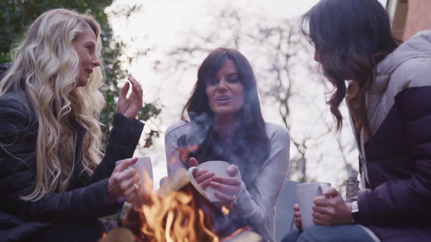 Three woman sit and talk with mugs by the campfire
