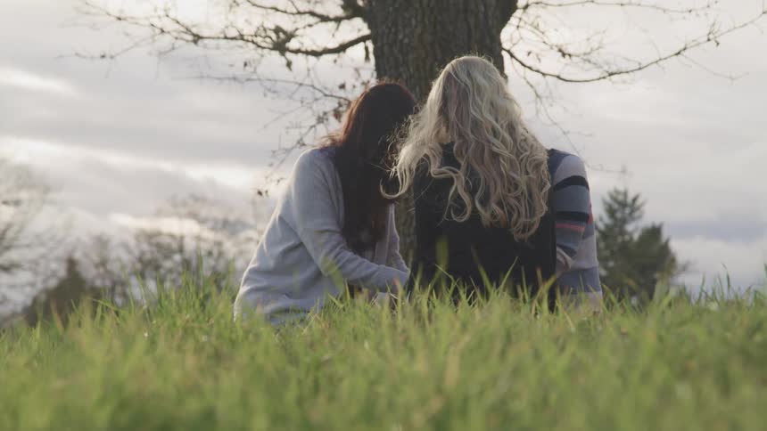 A group of women hold hands while sitting facing each other in a field on a cloudy day