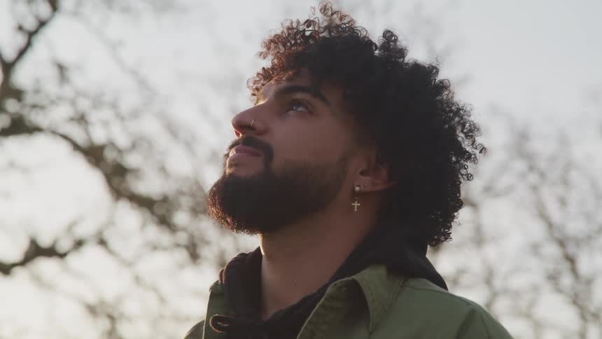 A young man standing in a grove of leafless trees looks upwards at the sky