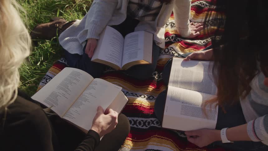 Three women are reading books while sitting on a blanket outdoors