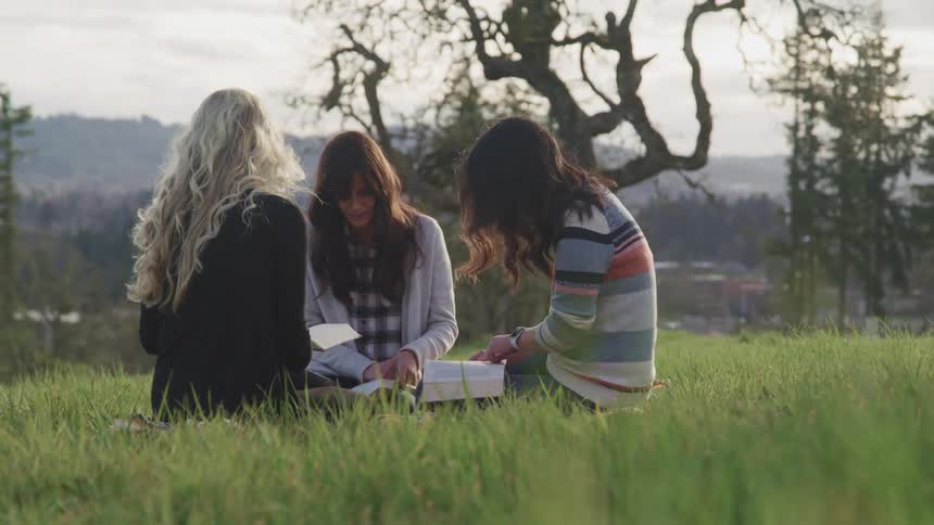 A group of female friends sits on green grass and reads books on a cloudy day