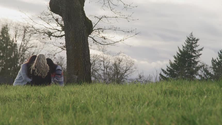 Three women sit beneath a tree holding hands in a circle and with their heads down