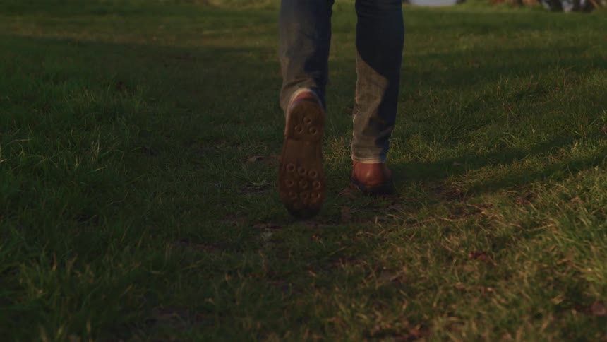 A person in black jeans and brown shoes walks through a field in the evening