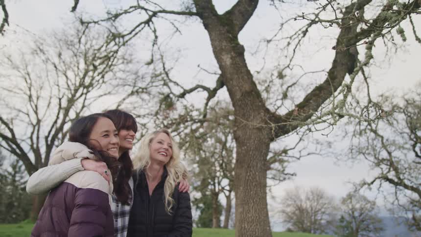Three women hug and laugh on a hill with trees