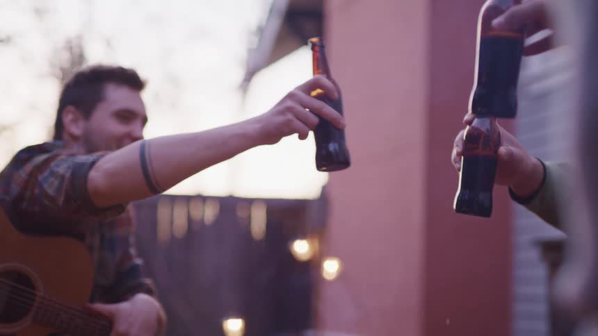 Men clink beer bottles smiling in backyard as smoke rises nearby