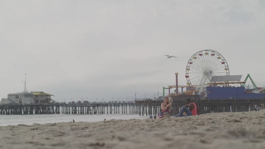 A man and woman sit on the beach by a pier and carnival