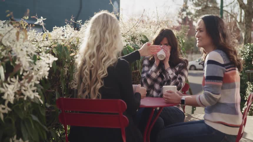 Three woman sit at a small outside table drinking from mugs and having a conversation