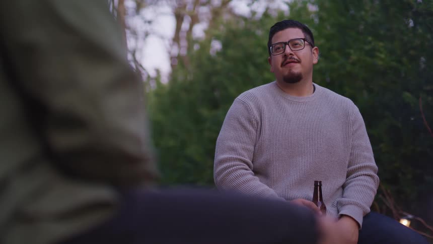A man with glasses holding a beer bottle sitting next to a campfire