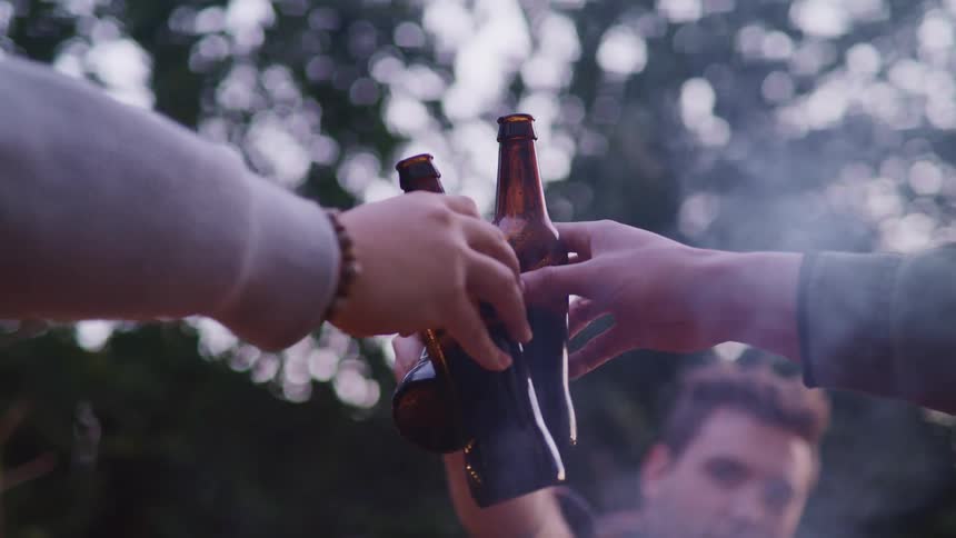 A group of men clink their beer bottles together while sitting around a campfire during the day