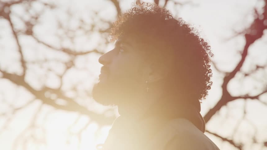 Side view of a man with curly hair a beard and cross earrings looking up as the sun shines brightly in the background