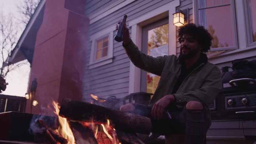 A man with a beer raised in the air sits outside a home around a firepit with another person