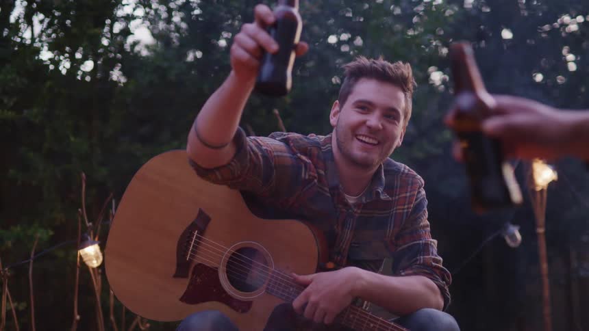 A group of people sit around a campfire and cheers their beer bottles together while one man holds a guitar