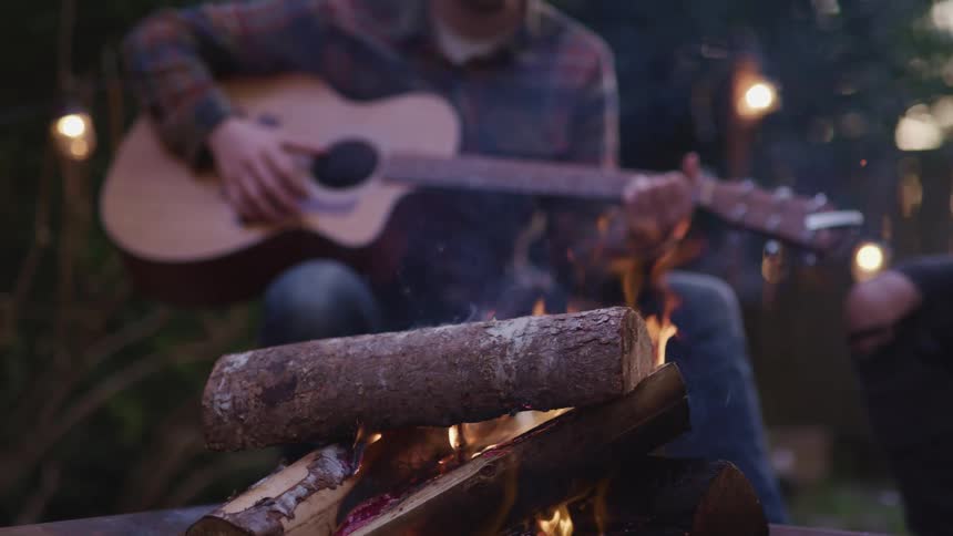 A man plays the guitar while sitting in front of a campfire