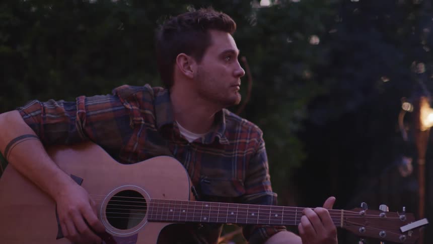 A man absentmindedly strums a guitar while sitting outdoors in the evening