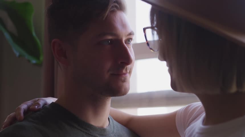 A man and a woman standing and hugging with a window and a plant in the background