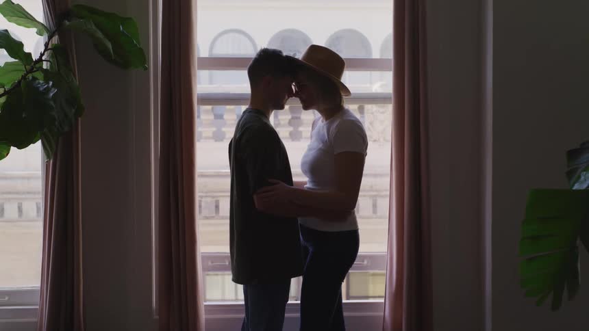 Young couple hug looking each other in the eyes in front of a window on a room