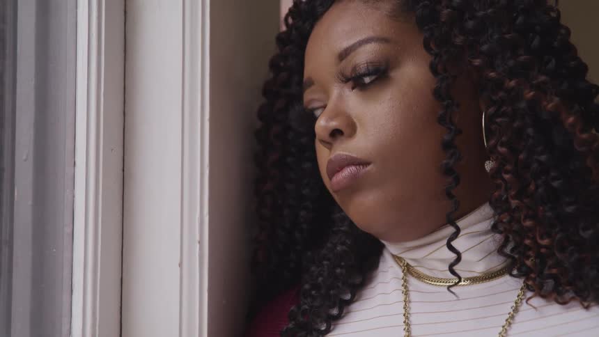 Black chubby woman with curly hair and hoop earrings looking out the windows feels bored and dull