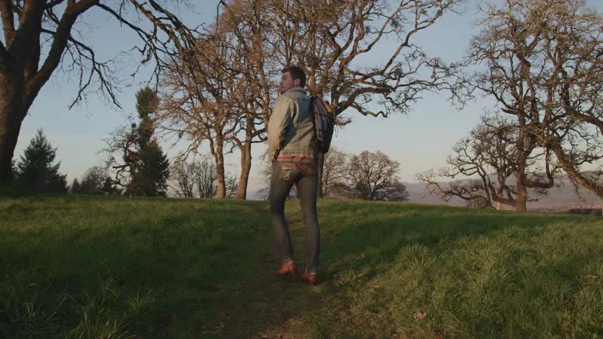 Man with jeans a denim jacket and a backpack walks on a dirt track at sunset