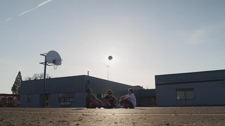 Three friends sit on the ground of an outdoor basketball court and throw the ball in the air