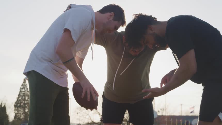 Men huddle while one holds a football under glaring sun
