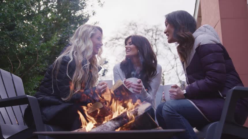 Group of friends talk to each other while sitting in the backyard in front of a fire drinking hot beverages