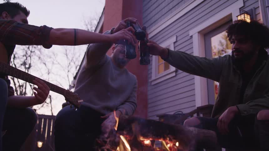 Three friends toast their beer bottles as they sit around a bonfire in their backyard