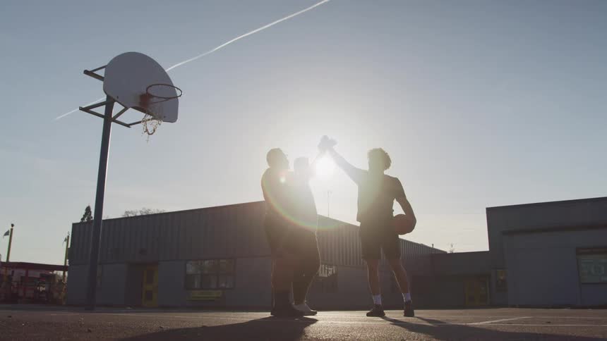 A group of men on an outdoor basketball court touch their fists together as the sun shines down on them