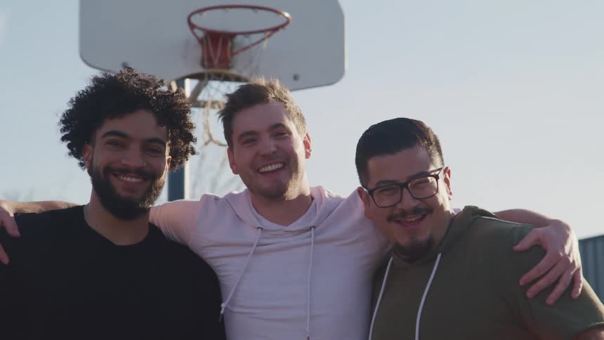 A man puts his arms around his two friends as they smile and pose in front of a basketball hoop