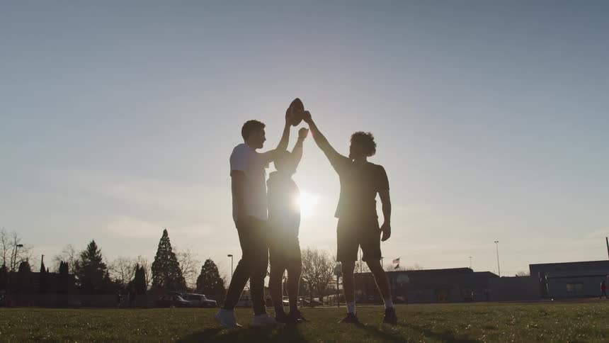 Three men in a field raise their arms to touch a football together as the sun shines down on the them