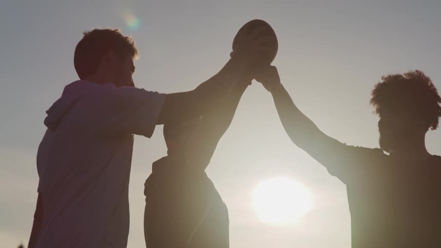 A group of young men raise their arms to touch a football together as the sun shines down on them