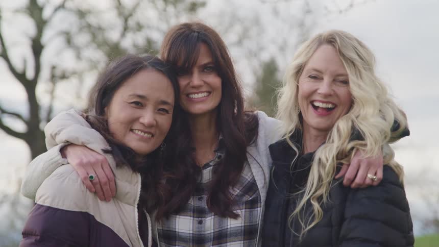 Three woman hugging and smiling with grass trees and the sky in the background