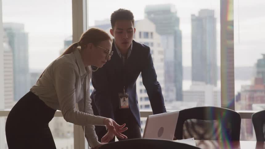 Two business people talk intently with someone on laptop while standing in conference room with view of city