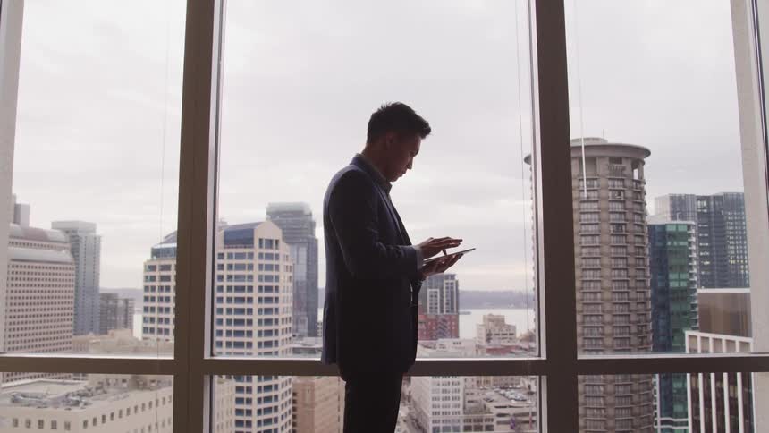 A man in a downtown office stands in front of a window and uses a tablet