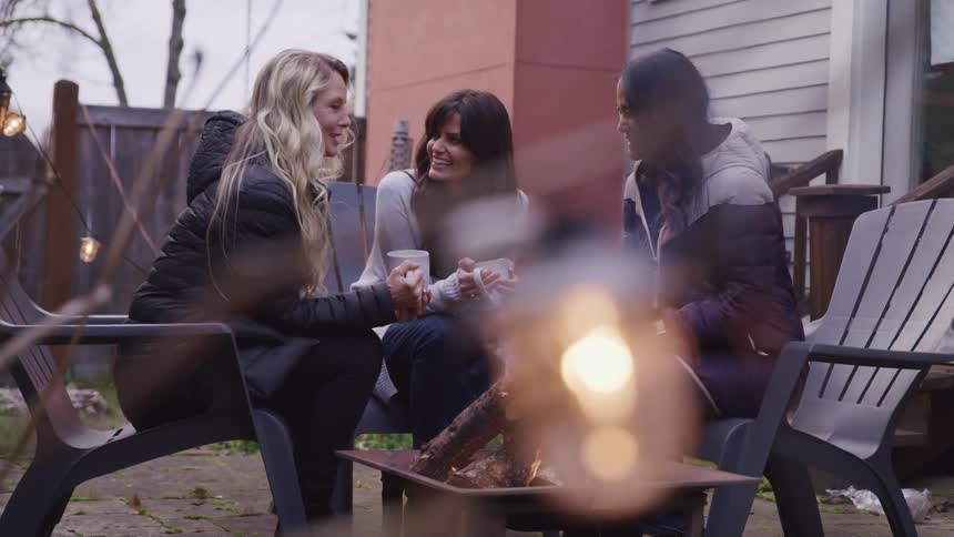 Three women sitting around a fire pit outside a home hold coffee cups and talk