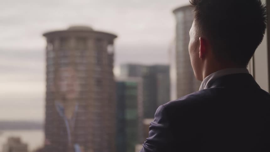 A man stands with his arms crossed and eyes closed in front of his office window with views of tall skyscrapers and downtown