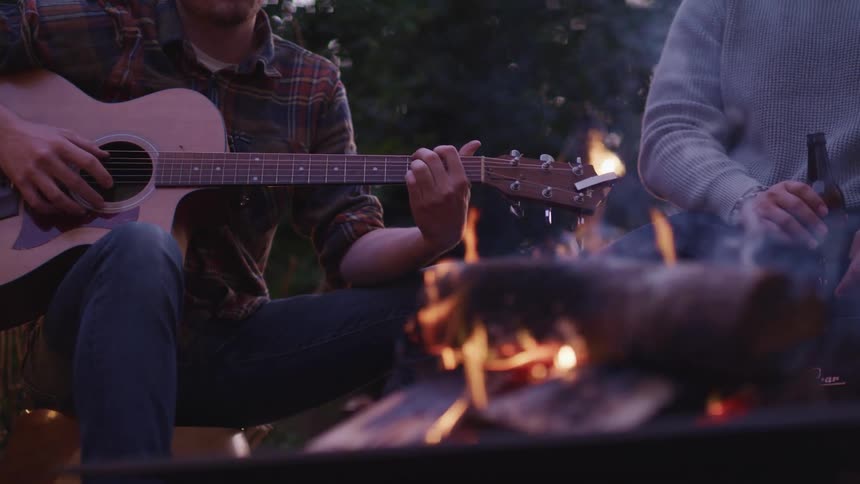 A man plays the guitar and another man drinks a beer as they sit around a fire pit