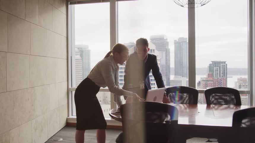 A man and a woman in a conference room looking at a laptop with a window and buildings in the background
