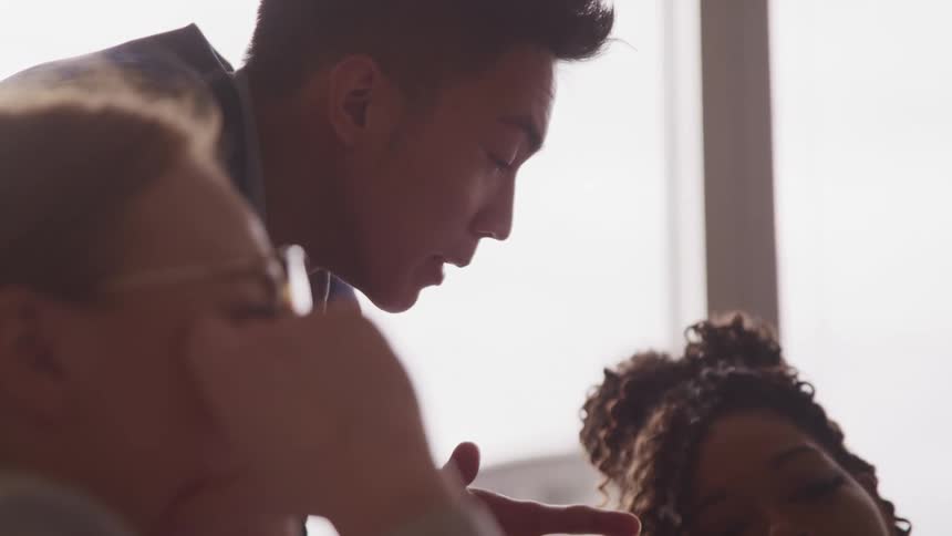 A young asian man in a suit talks to a pair of women sitting in an office