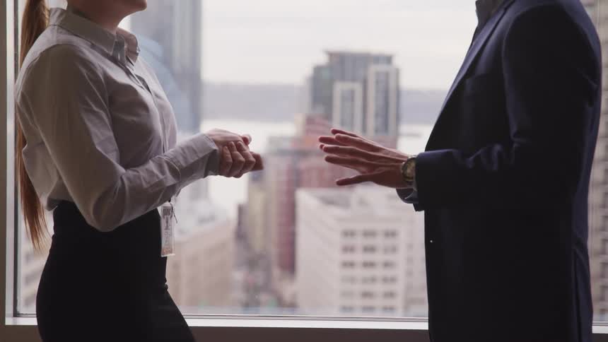 Man motions his hands while talking to a woman as they stand next to a window
