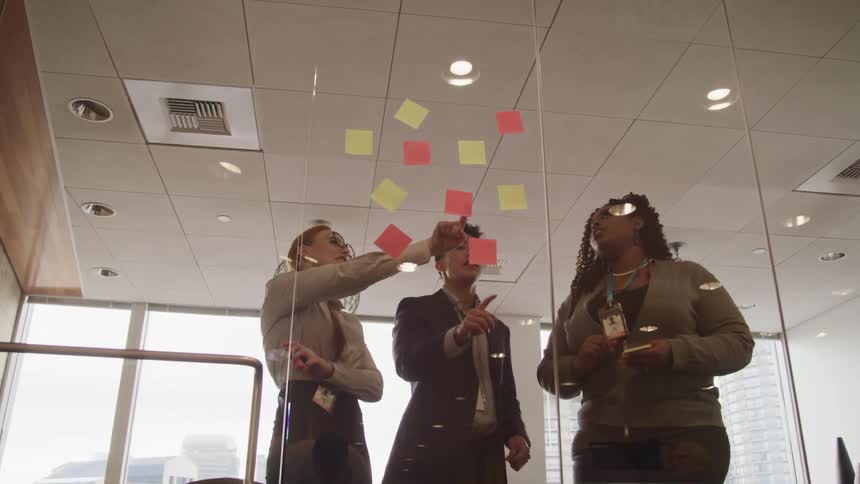 Three people gather in front of a glass office wall with sticky notes on it