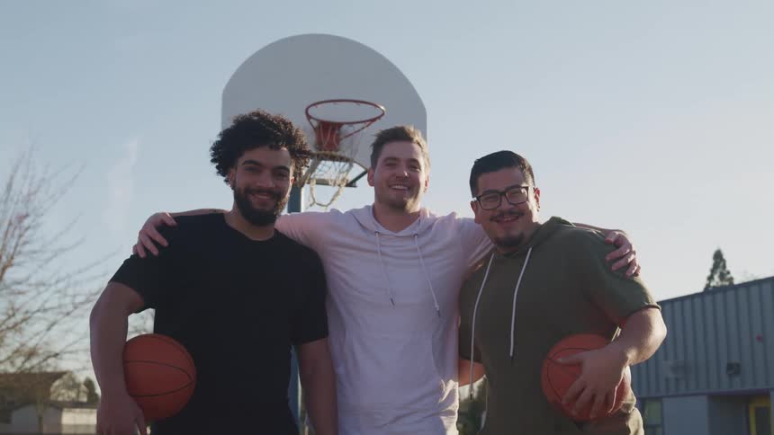 Three men pose together for a photo on an outdoors basketball court on a sunny day