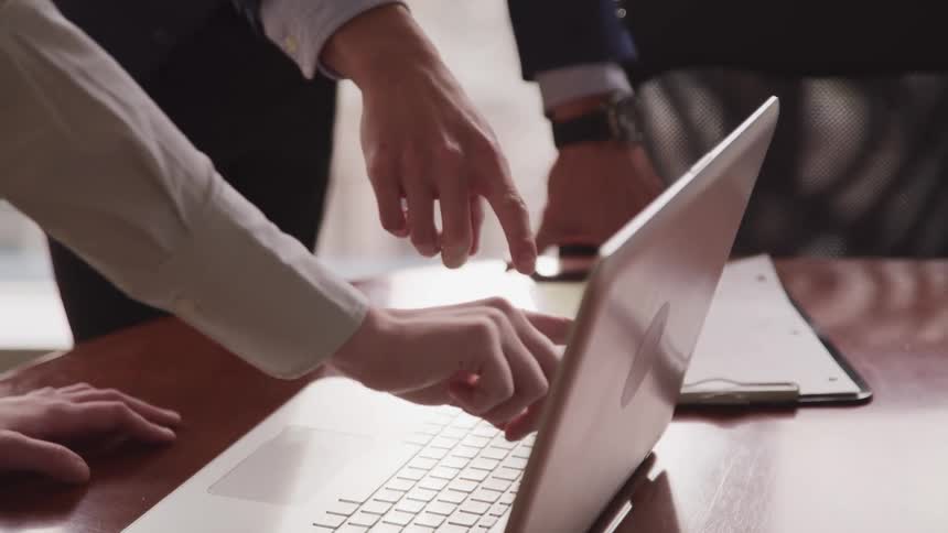 A pair of people point at the screen of a laptop as it rests on a wooden tabletop