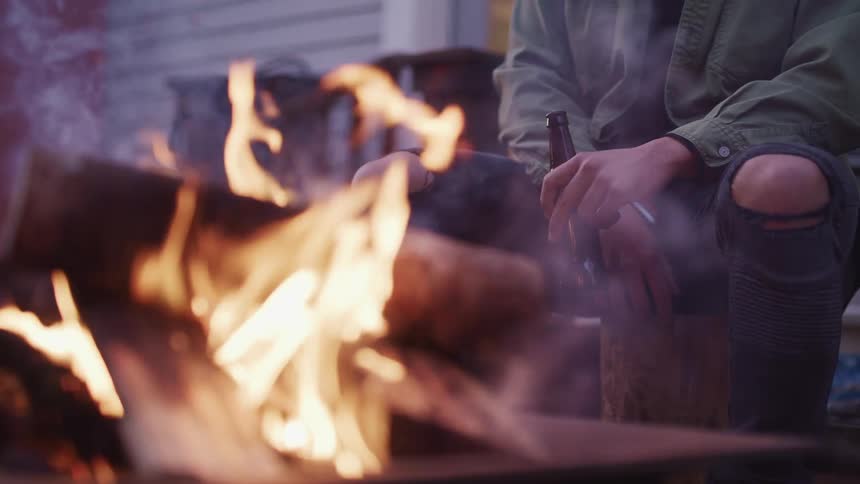 Young man with a beer bottle in hand sits in front of a campfire in the backyard